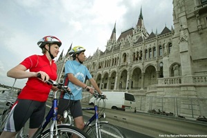 Ein Radlerpaar steht am Rand der Straße und blickt interessiert auf das Parlament von Budapest